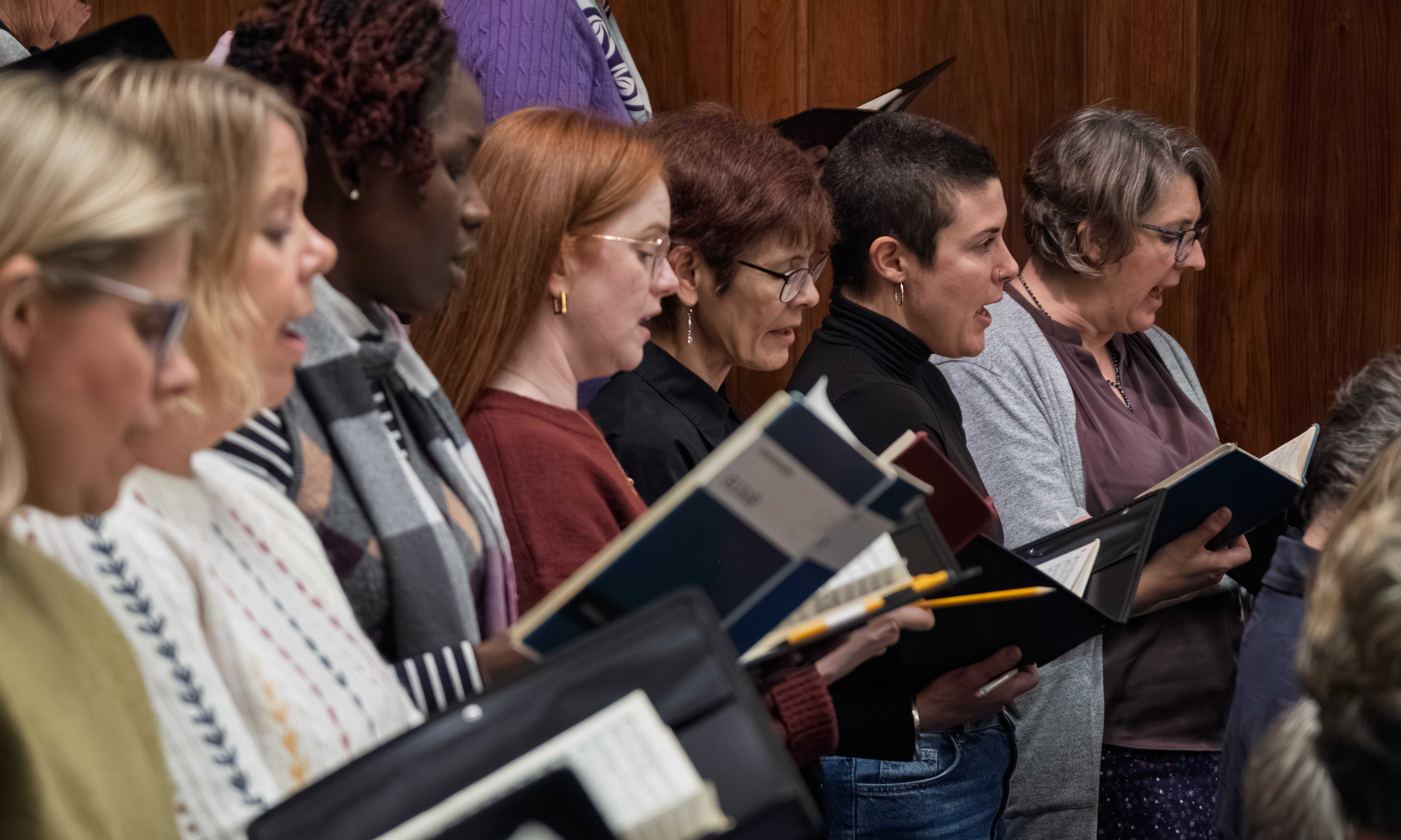 Close up of a group of choir members singing at a dress rehearsal.  They are all dressed informally and are holding musical scores.