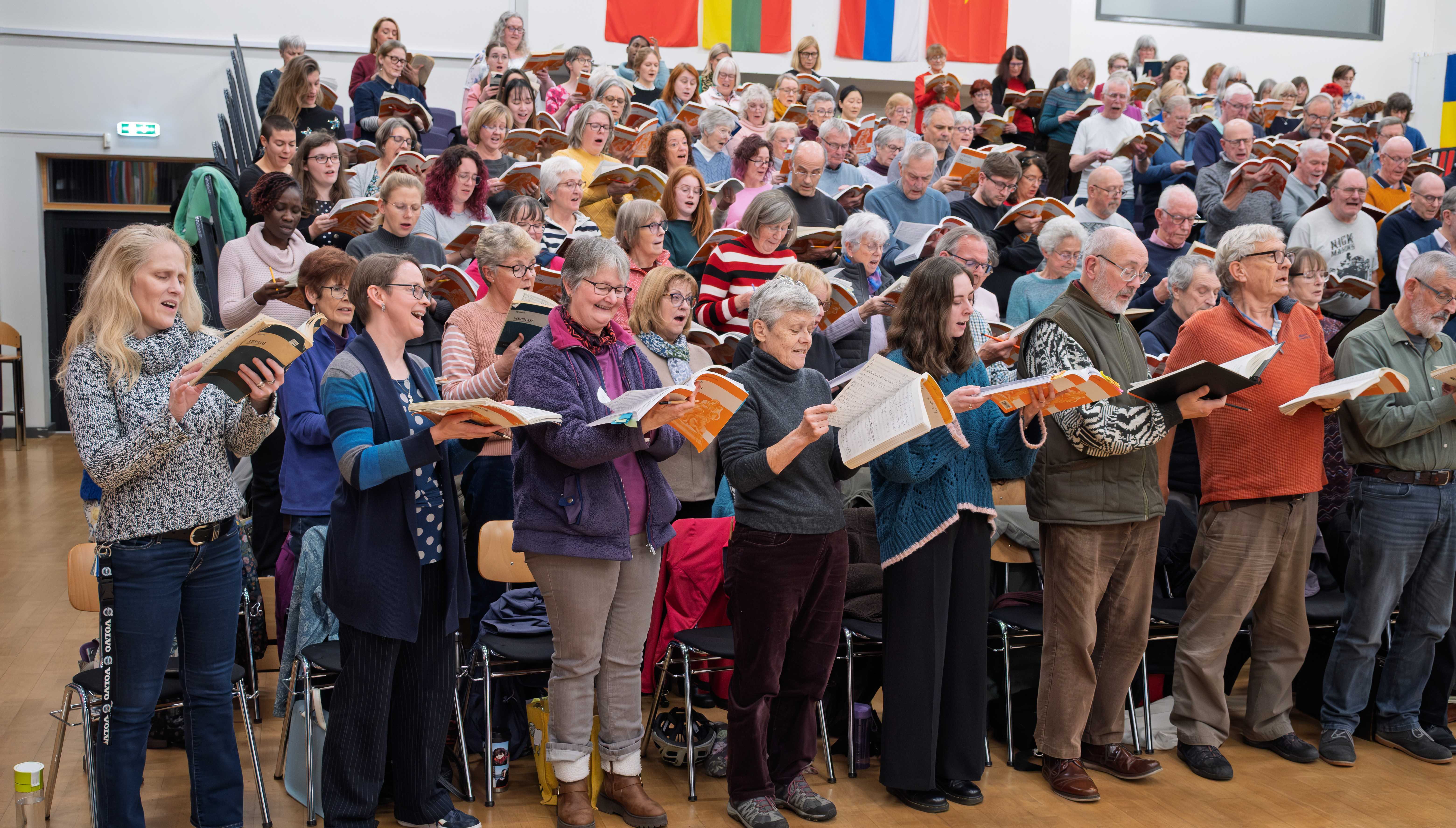 Part of the choir singing at a regular Monday night rehearsal.  The choir members are dressed informally and are holding their musical scores open in front of them.